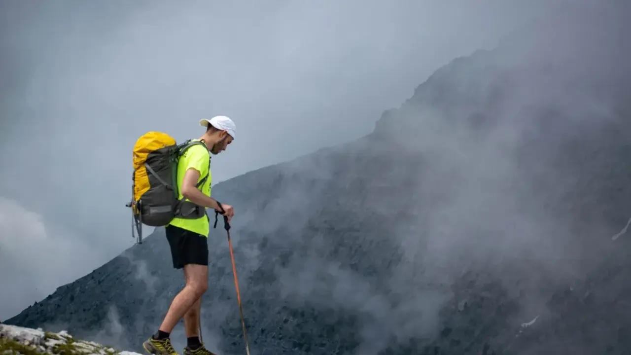 Mężczyzna z plecakiem i kijami trekkingowymi idzie po skalistym szlaku w górach, otoczony mgłą.