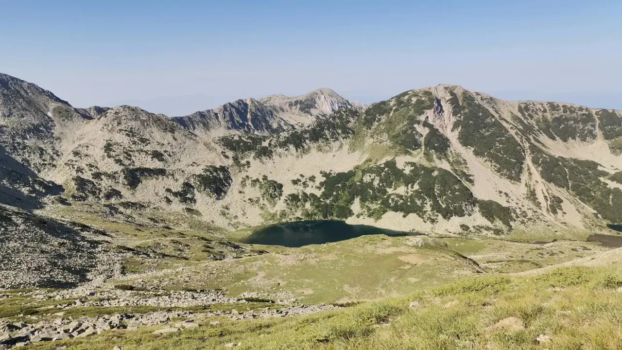 Widok na skaliste szczyty i zielone zbocza gór Pirin, z jeziorem w dolinie.