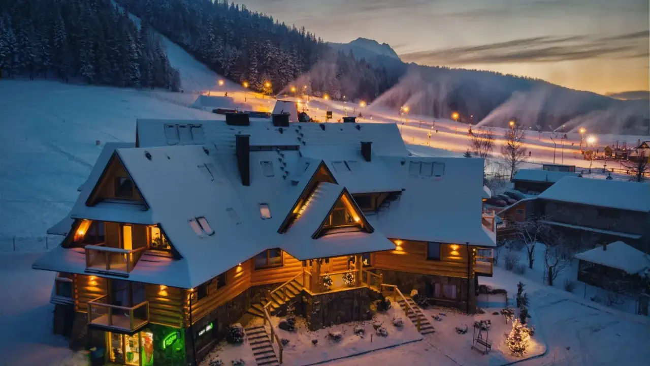 Drewniany pensjonat w śnieżnej scenerii, idealny na zakopane dla początkujących. Oświetlone stoki narciarskie w tle.
