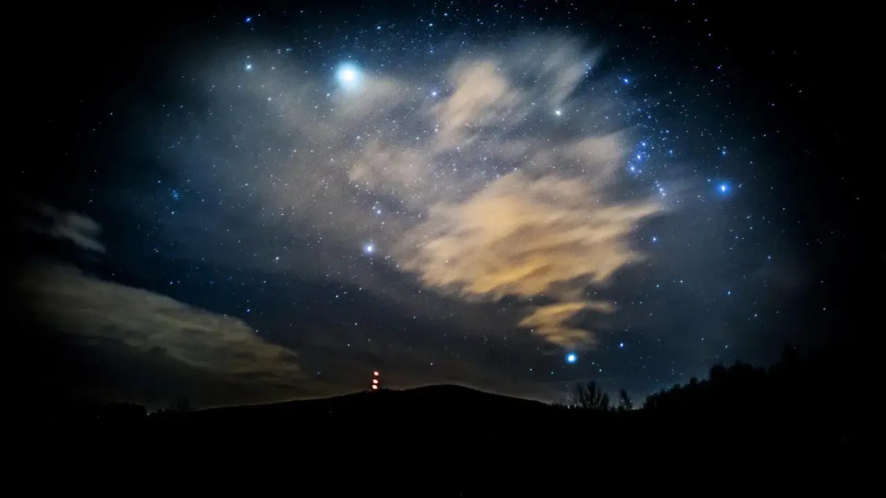 Nocne niebo pełne gwiazd, chmur i odległych świateł. W oddali widać zarys wzgórza z wieżą. To właśnie takie widoki towarzyszą nam, gdy zastanawiamy się, co to są Perseidy.