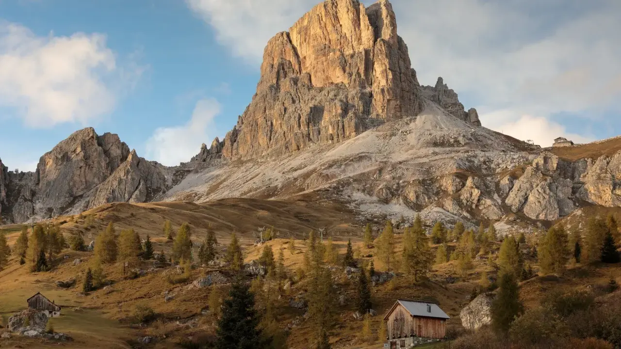 Jesienne słońce oświetla majestatyczne szczyty Dolomitów, idealne miejsce na trekking. Drewniana chata i drzewa tworzą malowniczy krajobraz.