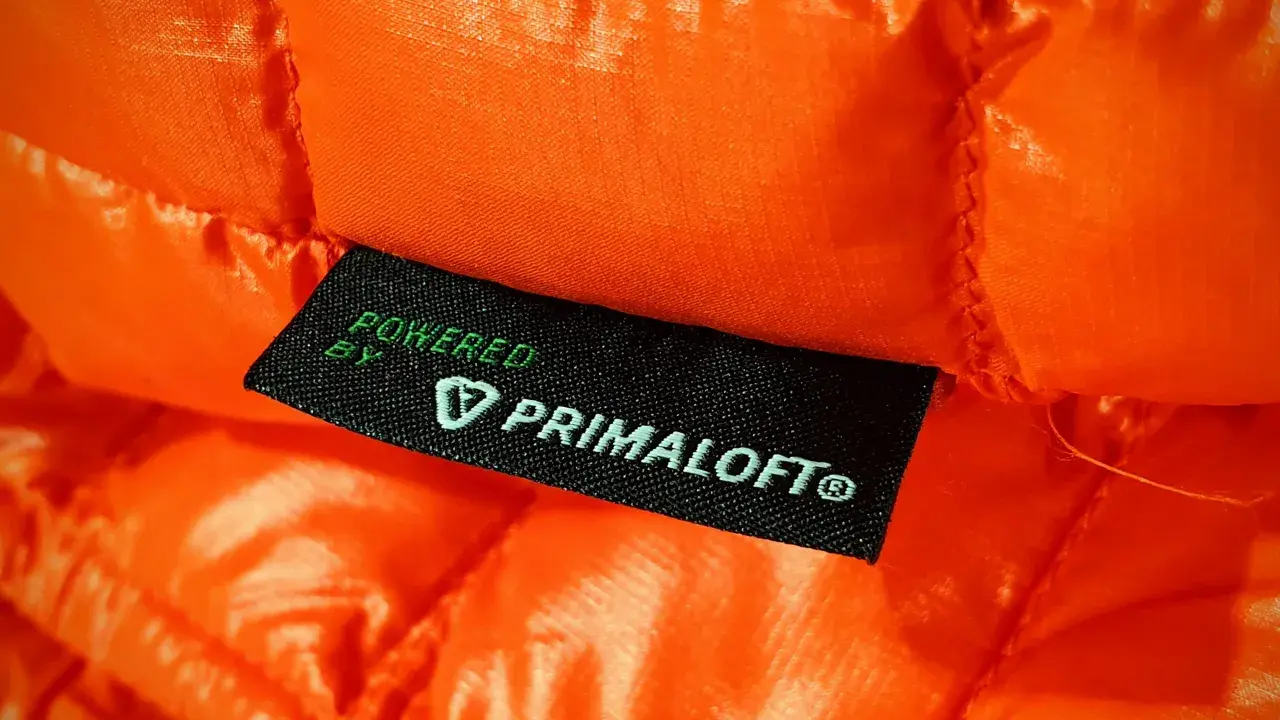 Pomarańczowa, pikowana tkanina z metką "Powered by Primaloft". To materiał izolacyjny, który zapewnia ciepło i lekkość.