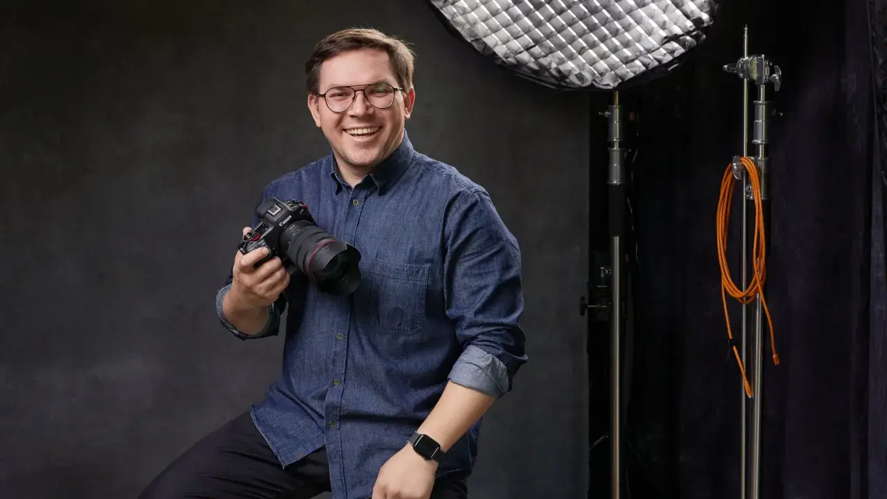 Uśmiechnięty mężczyzna z aparatem, gotowy poprowadzić kurs fotografii dla początkujących. W tle sprzęt studyjny.