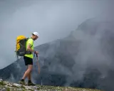 Mężczyzna z plecakiem i kijami trekkingowymi idzie po skalistym szlaku w górach, otoczony mgłą.