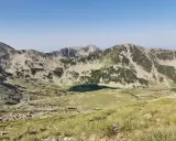 Widok na skaliste szczyty i zielone zbocza gór Pirin, z jeziorem w dolinie.