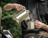 Mężczyzna nalewa kawę do kubka z metalowego czajniczka. Obok leży plecak i paczki kawy. Idealny **aeropress przepis** na kempingu.