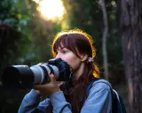 Młoda kobieta z aparatem w lesie, ćwiczy na zajęciach szkoły fotografii. Słońce przebija się przez drzewa.