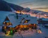 Drewniany pensjonat w śnieżnej scenerii, idealny na zakopane dla początkujących. Oświetlone stoki narciarskie w tle.