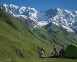 Para odpoczywa po trekkingu w Gruzji, siedząc obok zielonego namiotu na tle majestatycznych, ośnieżonych gór.
