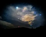 Nocne niebo pełne gwiazd, chmur i odległych świateł. W oddali widać zarys wzgórza z wieżą. To właśnie takie widoki towarzyszą nam, gdy zastanawiamy się, co to są Perseidy.