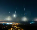 Noc perseidów nad miastem. Spadające gwiazdy przecinają ciemne niebo, oświetlając mgliste doliny i rozświetlone światłami zabudowania.
