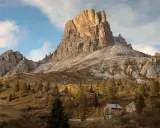 Jesienne słońce oświetla majestatyczne szczyty Dolomitów, idealne miejsce na trekking. Drewniana chata i drzewa tworzą malowniczy krajobraz.