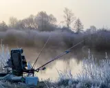 Mroźny poranek nad rzeką. Krzesło wędkarskie, wędki i sprzęt czekają na ryby. W tle mgła i oszronione drzewa.
