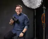 Uśmiechnięty mężczyzna z aparatem, gotowy poprowadzić kurs fotografii dla początkujących. W tle sprzęt studyjny.