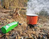 Jaka kuchenka turystyczna! Zielony kartusz z gazem i czajnik na palniku, parujący nad wodą w lesie.