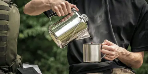 Mężczyzna nalewa kawę do kubka z metalowego czajniczka. Obok leży plecak i paczki kawy. Idealny **aeropress przepis** na kempingu.