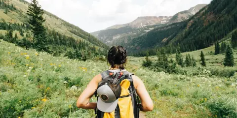 Kobieta z plecakiem wyrusza na trekking przez kwiecistą łąkę w górach.