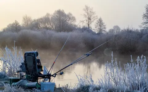 Mroźny poranek nad rzeką. Krzesło wędkarskie, wędki i sprzęt czekają na ryby. W tle mgła i oszronione drzewa.
