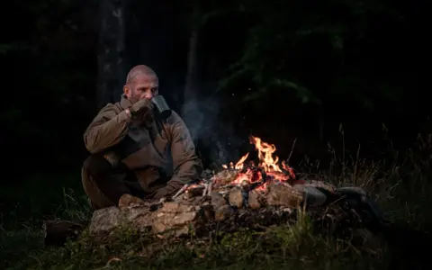 Mężczyzna pije gorący napój przy ognisku, przygotowując się na burzę w górach. Dym unosi się w powietrzu, a płomienie tańczą na kamieniach.
