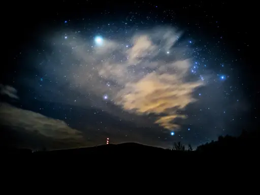 Nocne niebo pełne gwiazd, chmur i odległych świateł. W oddali widać zarys wzgórza z wieżą. To właśnie takie widoki towarzyszą nam, gdy zastanawiamy się, co to są Perseidy.