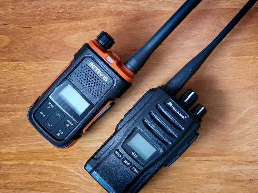 Dwa radiotelefony, jeden pomarańczowo-czarny RETC/IS, drugi czarny Midland G13, leżą na drewnianym blacie.