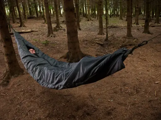 Mężczyzna śpi w czarnym śpiworze zawieszonym między drzewami w lesie. Spanie w hamaku to przygoda.