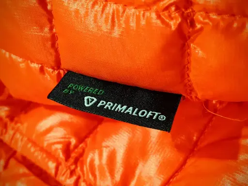 Pomarańczowa, pikowana tkanina z metką "Powered by Primaloft". To materiał izolacyjny, który zapewnia ciepło i lekkość.