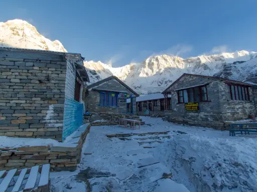 Kamienne budynki na trasie Annapurna Trekking, pokryte śniegiem, z majestatycznymi, ośnieżonymi szczytami w tle.