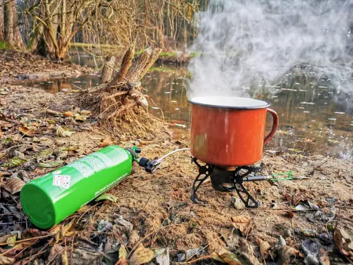 Jaka kuchenka turystyczna! Zielony kartusz z gazem i czajnik na palniku, parujący nad wodą w lesie.