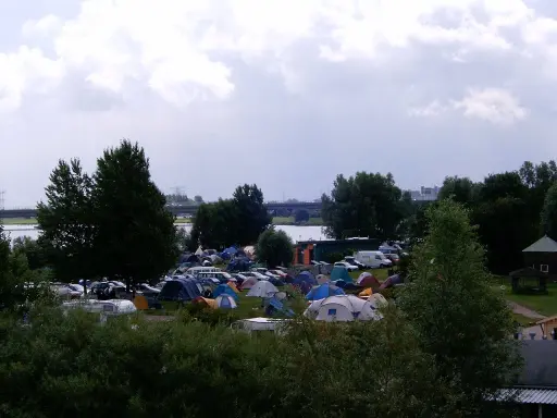 Rozległy kamping z mnóstwem namiotów i samochodów, nad wodą, pod pochmurnym niebem.
