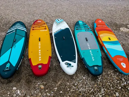 Pięć desek SUP leży na plaży. Zastanawiasz się, **deska sup jaką wybrać**? Wybierz swoją idealną!
