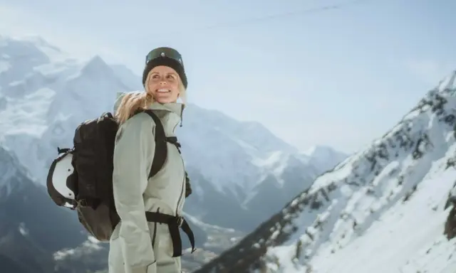 Kobieta w narciarskim stroju z plecakiem, do którego przymocowany jest kask, na tle gór.