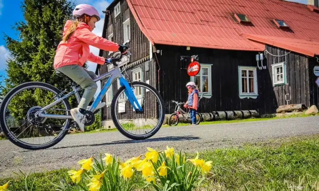 Dwie dziewczynki na rowerach cieszą się wiosennym dniem w czeskich Izera. Przed nimi kwitnące żonkile i drewniany domek.