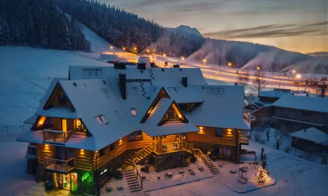 Drewniany pensjonat w śnieżnej scenerii, idealny na zakopane dla początkujących. Oświetlone stoki narciarskie w tle.