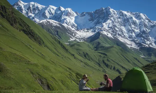 Para odpoczywa po trekkingu w Gruzji, siedząc obok zielonego namiotu na tle majestatycznych, ośnieżonych gór.