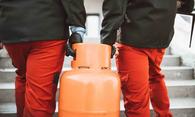 Dwóch mężczyzn w pomarańczowych spodniach niesie butlę z gazem. Wygląda na to, że nie można zakręcić butli z gazem, co może być problemem.