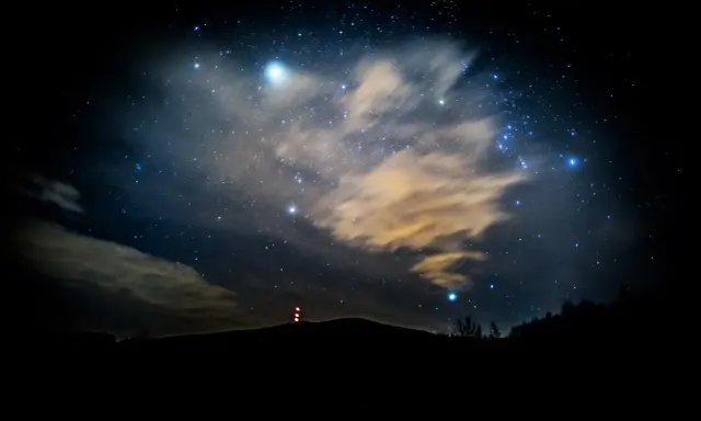 Nocne niebo pełne gwiazd, chmur i odległych świateł. W oddali widać zarys wzgórza z wieżą. To właśnie takie widoki towarzyszą nam, gdy zastanawiamy się, co to są Perseidy.