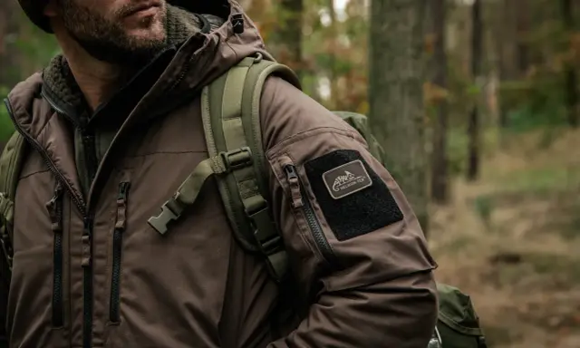Mężczyzna w brązowej kurtce z naszywką Helikon-Tex, symbol polskie marki sportowe, w lesie.