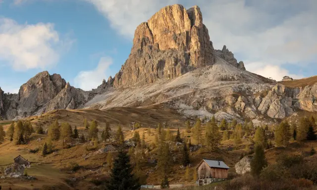 Jesienne słońce oświetla majestatyczne szczyty Dolomitów, idealne miejsce na trekking. Drewniana chata i drzewa tworzą malowniczy krajobraz.