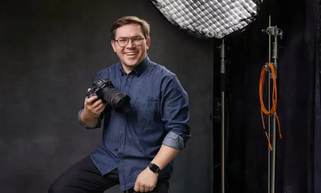 Uśmiechnięty mężczyzna z aparatem, gotowy poprowadzić kurs fotografii dla początkujących. W tle sprzęt studyjny.