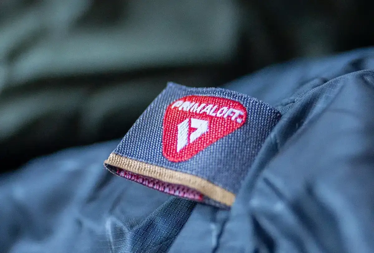 Metka z logo Primaloft na ciemnym materiale. Primaloft to wysokiej jakości izolacja, która zapewnia ciepło i komfort.