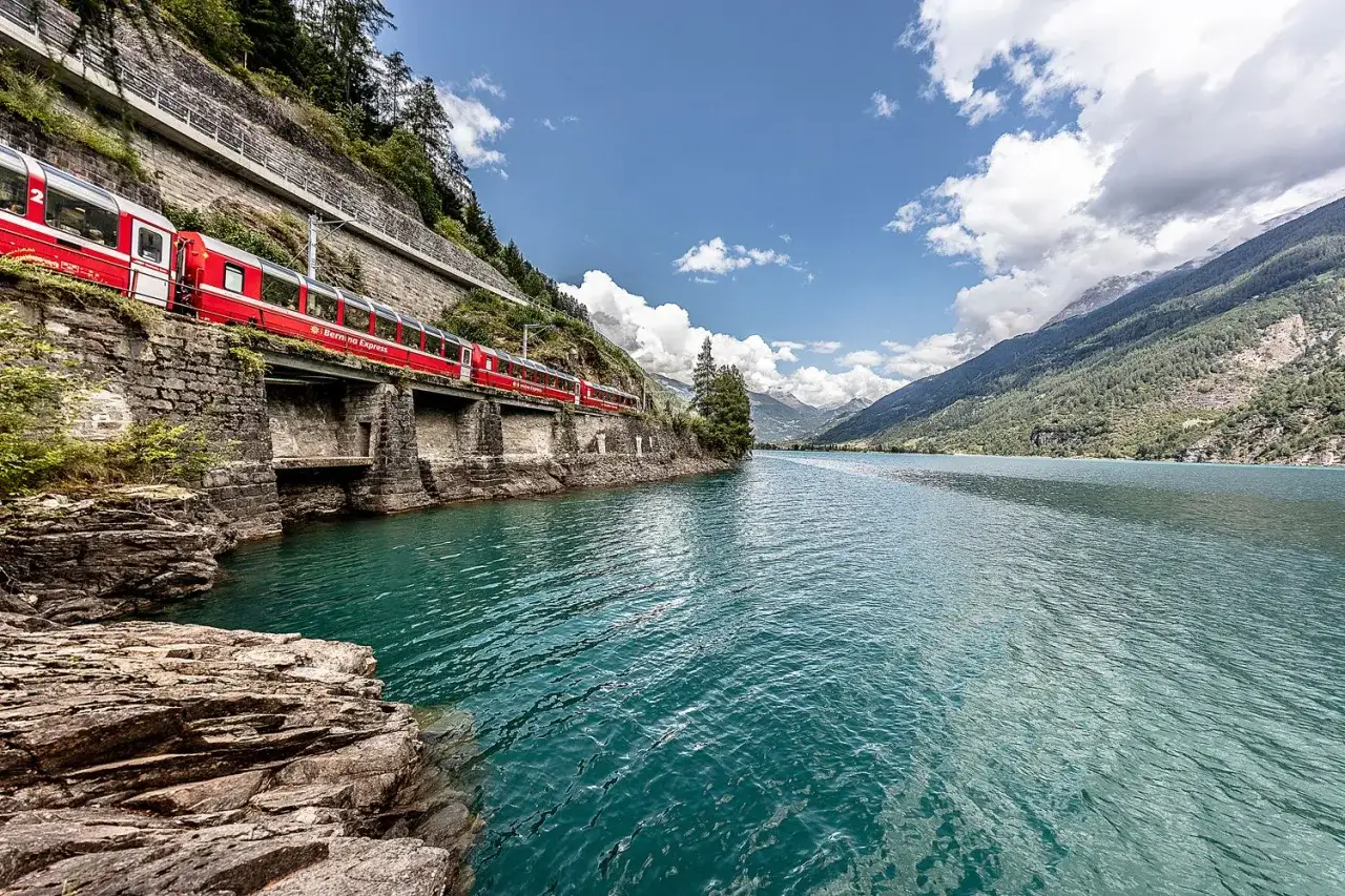Czerwony pociąg Bernina Express jedzie wzdłuż turkusowego jeziora, idealny widok dla posiadaczy Swiss Travel Pass.