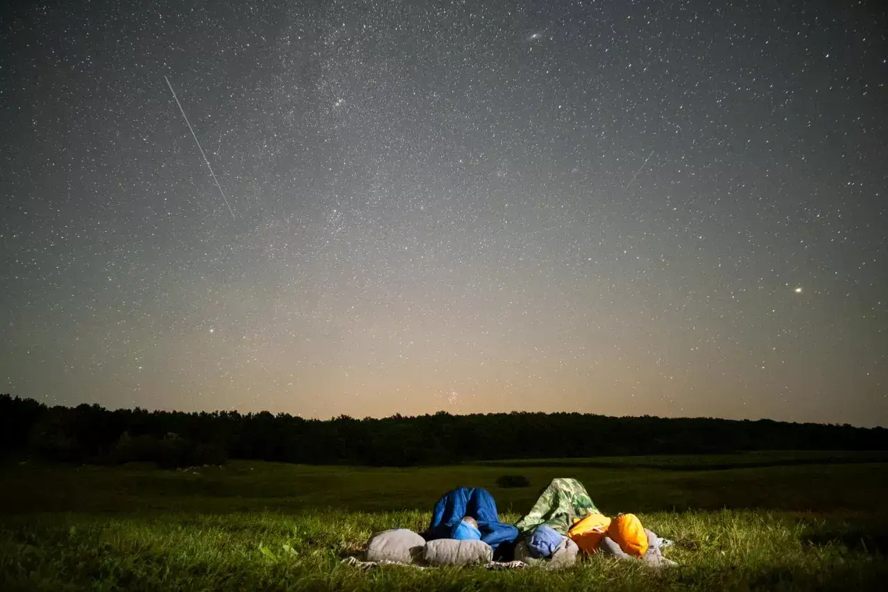 Nocne niebo pełne gwiazd, a na pierwszym planie śpiwory i plecaki. Widać też smugę spadającej gwiazdy – to właśnie co to są Perseidy, czyli deszcz meteorów.