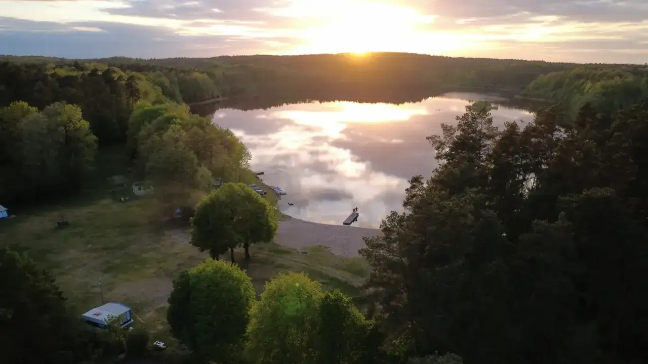 Zachód słońca nad jeziorem, odbijającym się w spokojnej tafli wody. Na brzegu plaża, kilka łódek i pomost, na którym stoją dwie osoby.