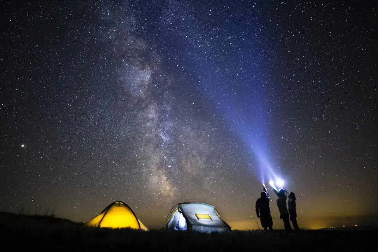 Nocne biwakowanie pod rozgwieżdżonym niebem. Dwa namioty i ludzie oświetlający latarkami Drogę Mleczną.