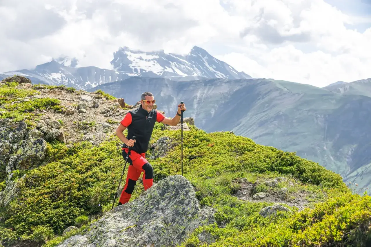 Mężczyzna w czerwonych spodniach i kamizelce, z okularami przeciwsłonecznymi, wspina się po skalistym zboczu, używając kijków trekkingowych. W tle majestatyczne, ośnieżone góry.