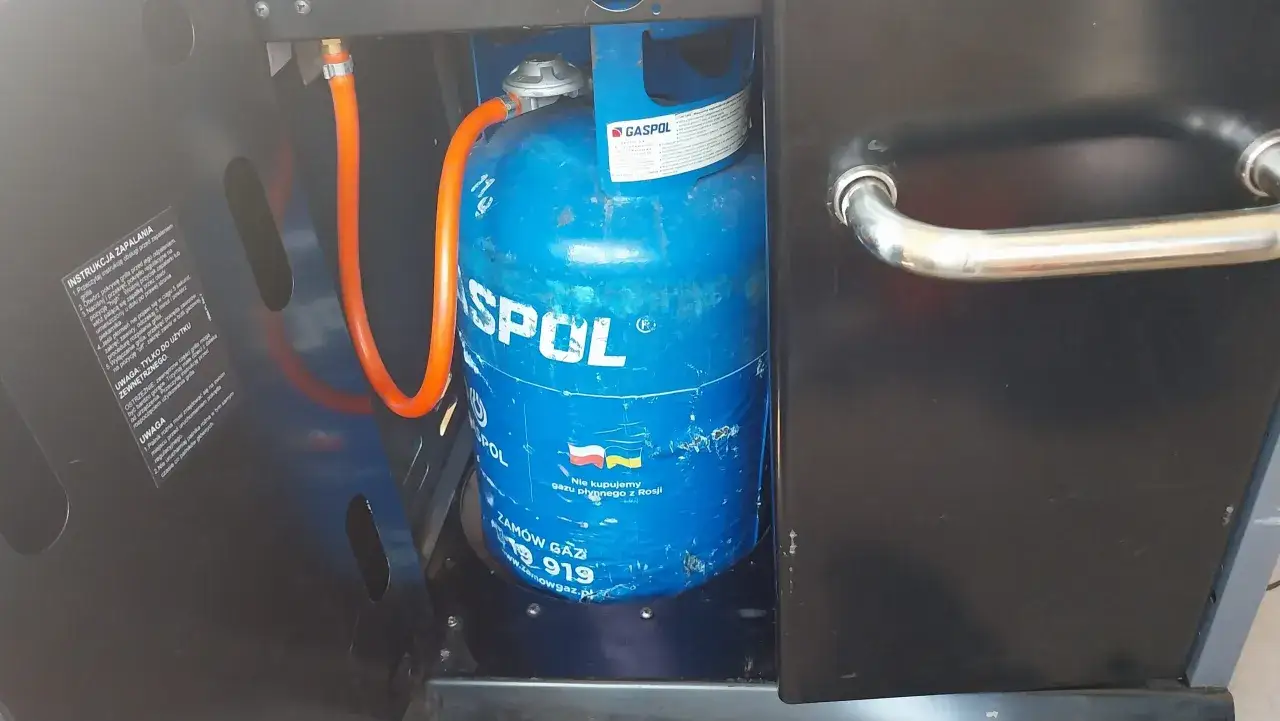 Błękitna butla z gazem marki GASPOL, podłączona pomarańczowym wężem, stanowi serce grilla. To idealna butla do grilla gazowego, gotowa do akcji.
