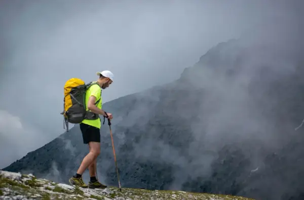 Mężczyzna z plecakiem i kijami trekkingowymi idzie po skalistym szlaku w górach, otoczony mgłą.