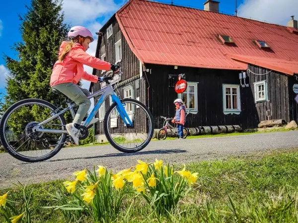 Dwie dziewczynki na rowerach cieszą się wiosennym dniem w czeskich Izera. Przed nimi kwitnące żonkile i drewniany domek.