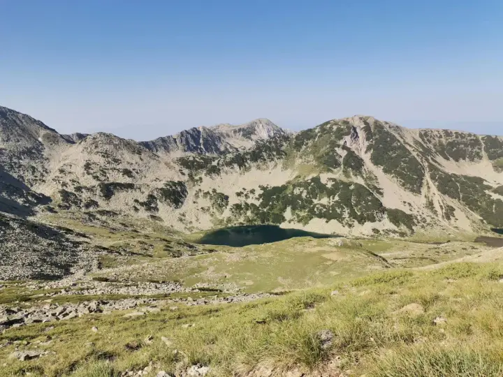 Widok na skaliste szczyty i zielone zbocza gór Pirin, z jeziorem w dolinie.